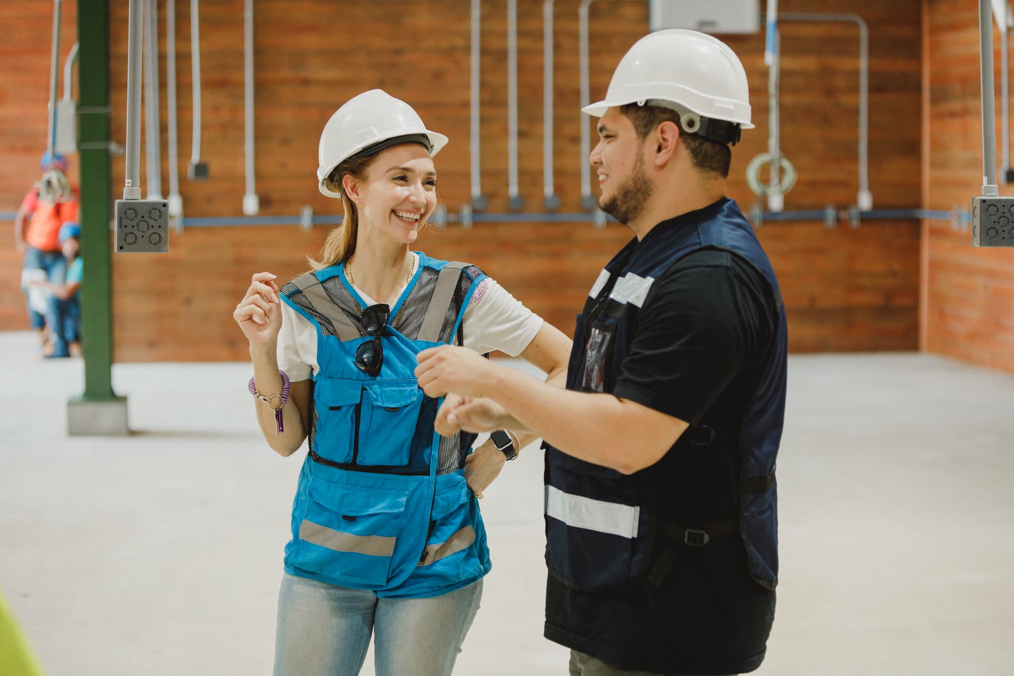 Two construction workers wearing hard hats and safety vests, engaged in a conversation inside a building under construction. They are smiling and appear to be discussing something related to their work, creating a positive and collaborative atmosphere.