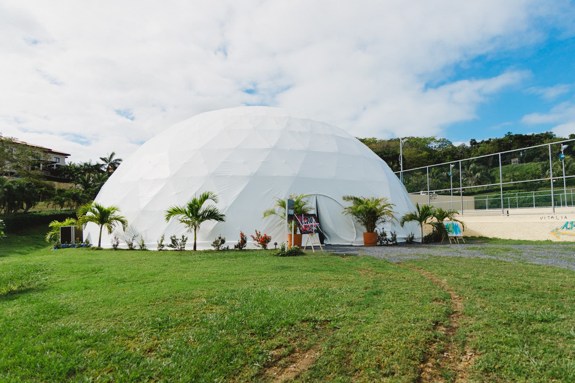 A large white geodesic dome set up on a grassy area, with tropical plants and small trees around the entrance. The dome is likely used for events or gatherings, and the sky above is partly cloudy, creating a bright and airy atmosphere.