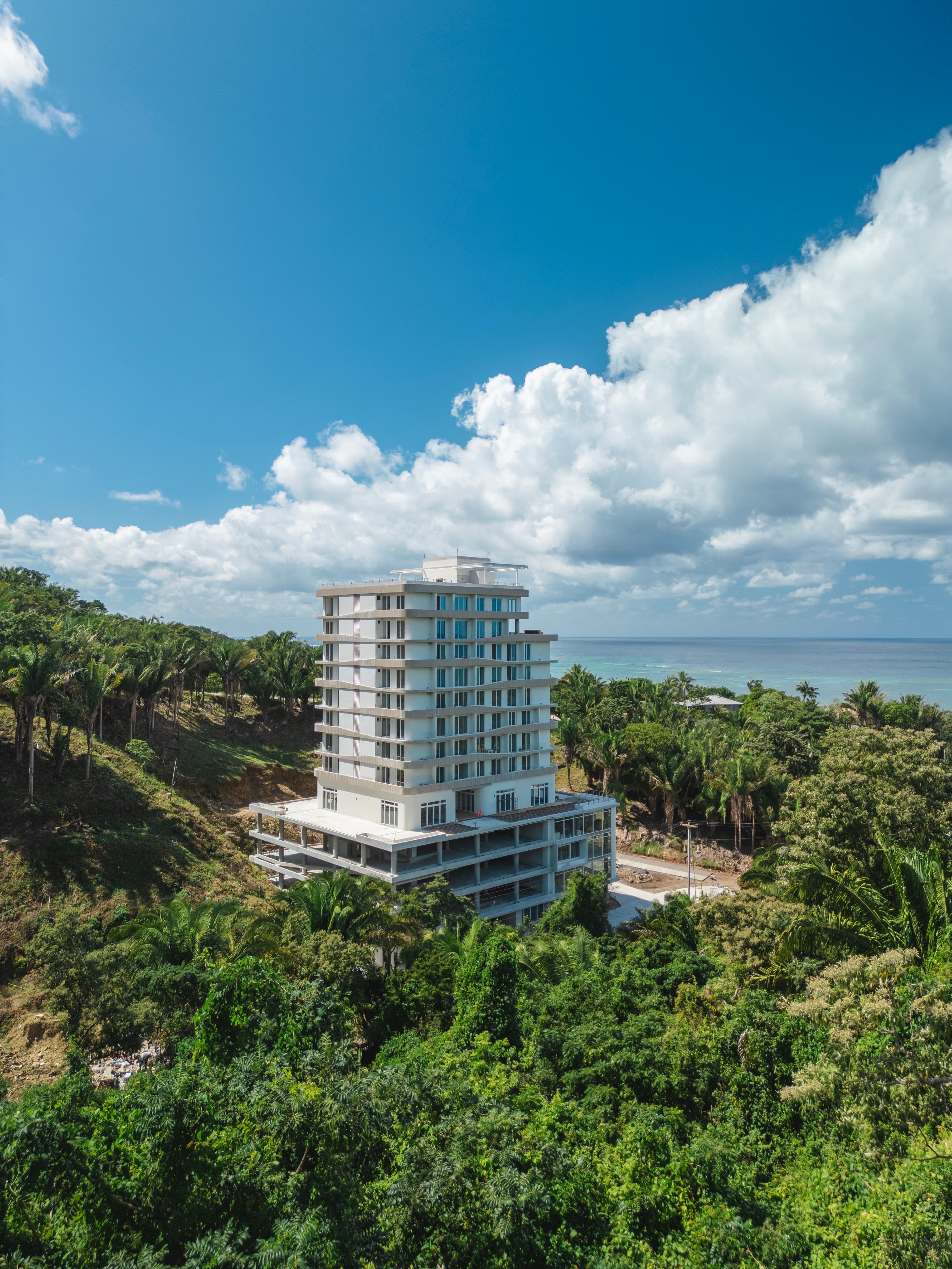 A tall, modern building under construction surrounded by dense greenery and palm trees, with the ocean visible in the background. The structure stands out against the blue sky with scattered clouds, indicating a coastal location with a mix of natural beauty and developing infrastructure.