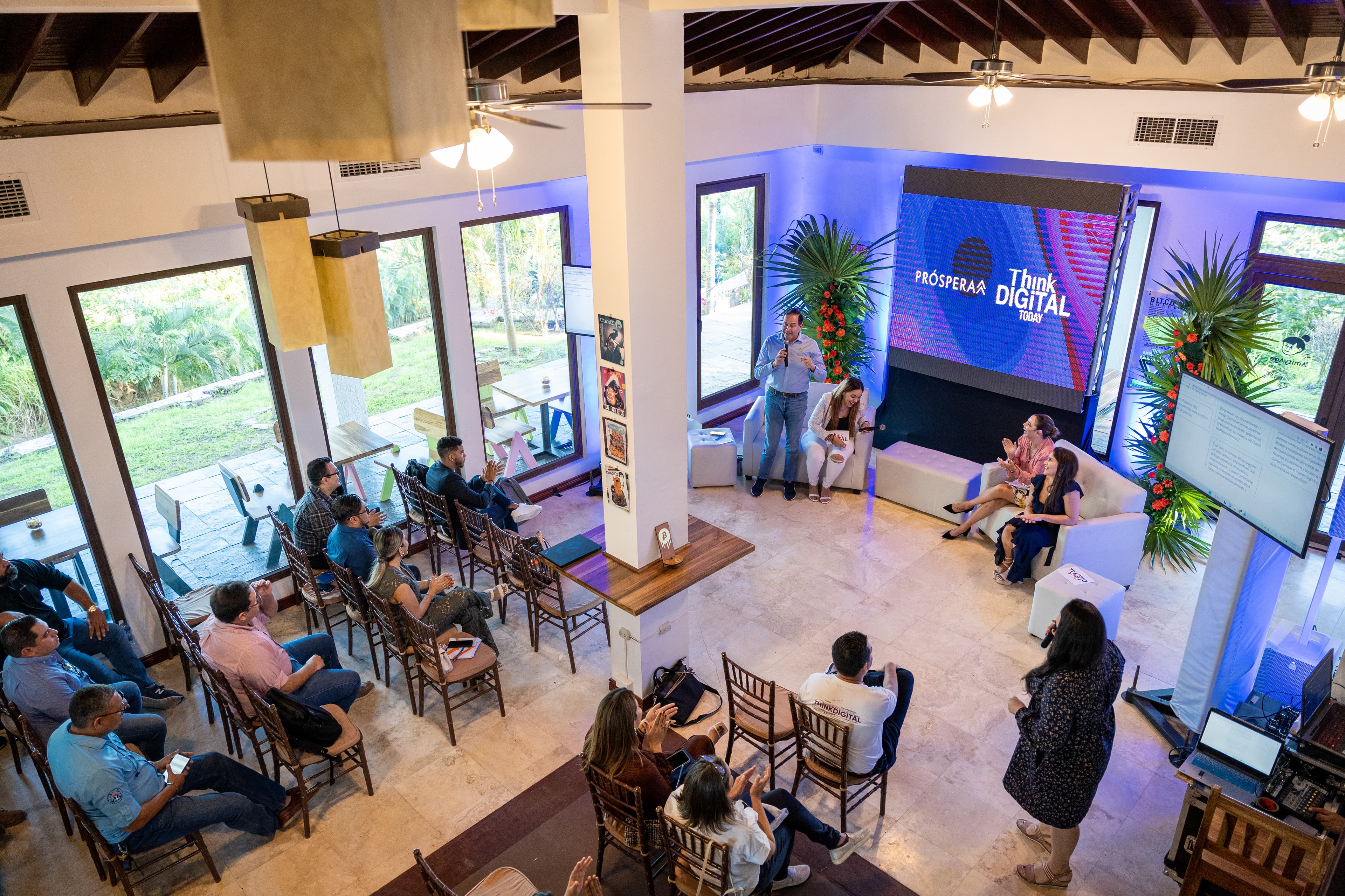 Indoor conference or seminar with attendees seated in rows, listening to speakers on a stage. A large screen behind the speakers displays PROSPERA and Think Digital, with plants and colorful decorations enhancing the setting. The room has large windows allowing natural light to brighten the space.