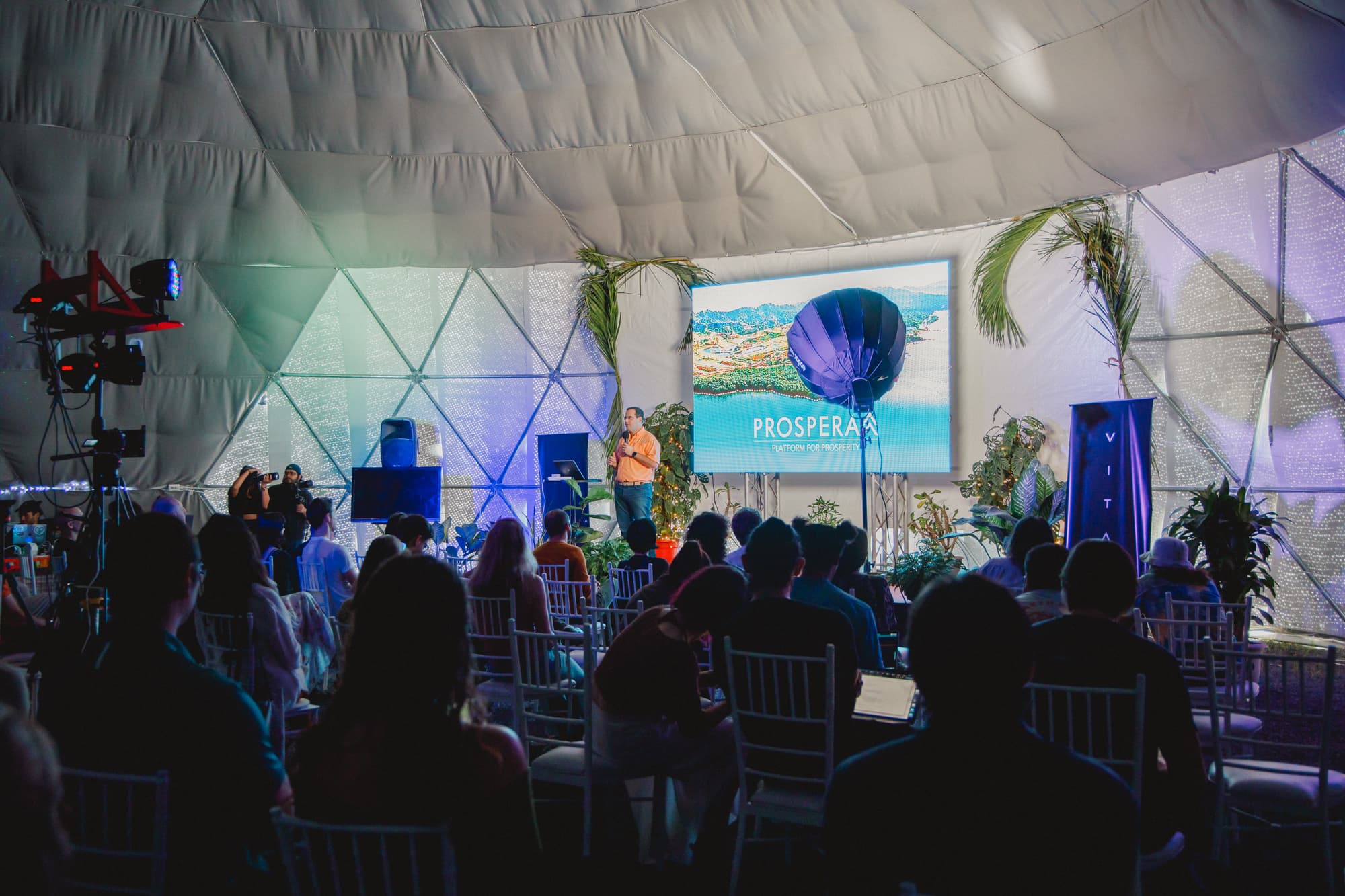 A man speaking on stage inside a geodesic dome, with a large screen displaying PROSPERA and Platform for Prosperity alongside an aerial image. The audience is seated in front, and stage lighting equipment is visible to the left.