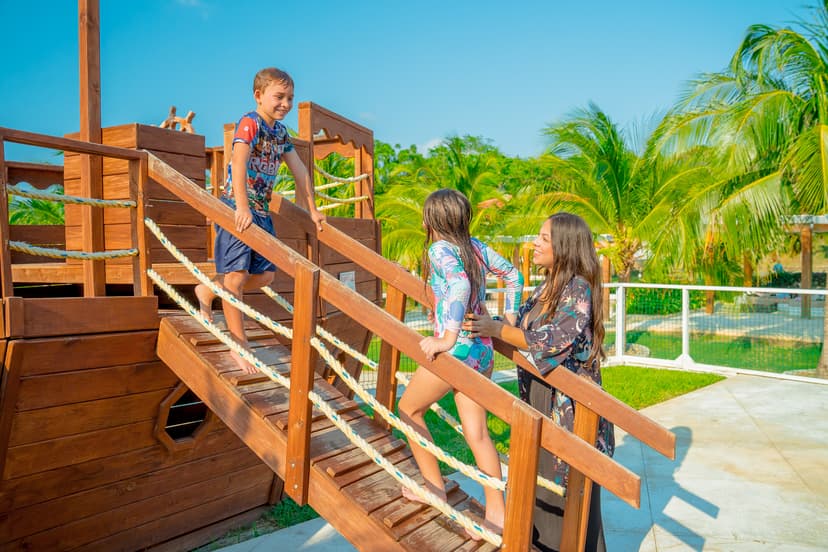 A boy standing on the wooden bridge of a play structure, smiling down at a girl and a woman encouraging him. The play area is set outdoors with palm trees and a clear blue sky in the background.