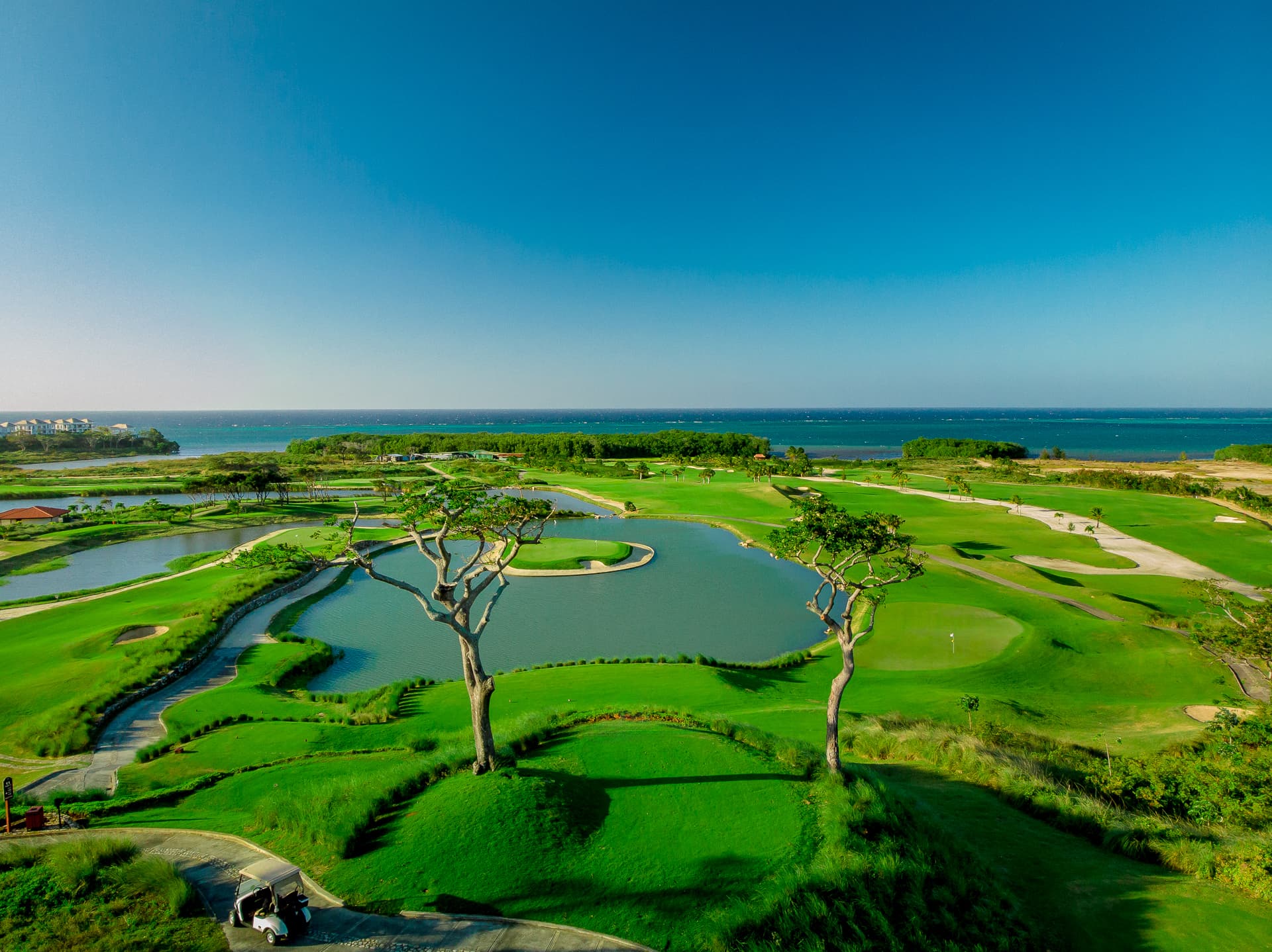 Aerial view of a golf course featuring well-maintained greens, sand traps, and several water hazards, with the ocean visible in the distance. The course is lush and green, set against a clear blue sky, creating a serene and picturesque landscape.