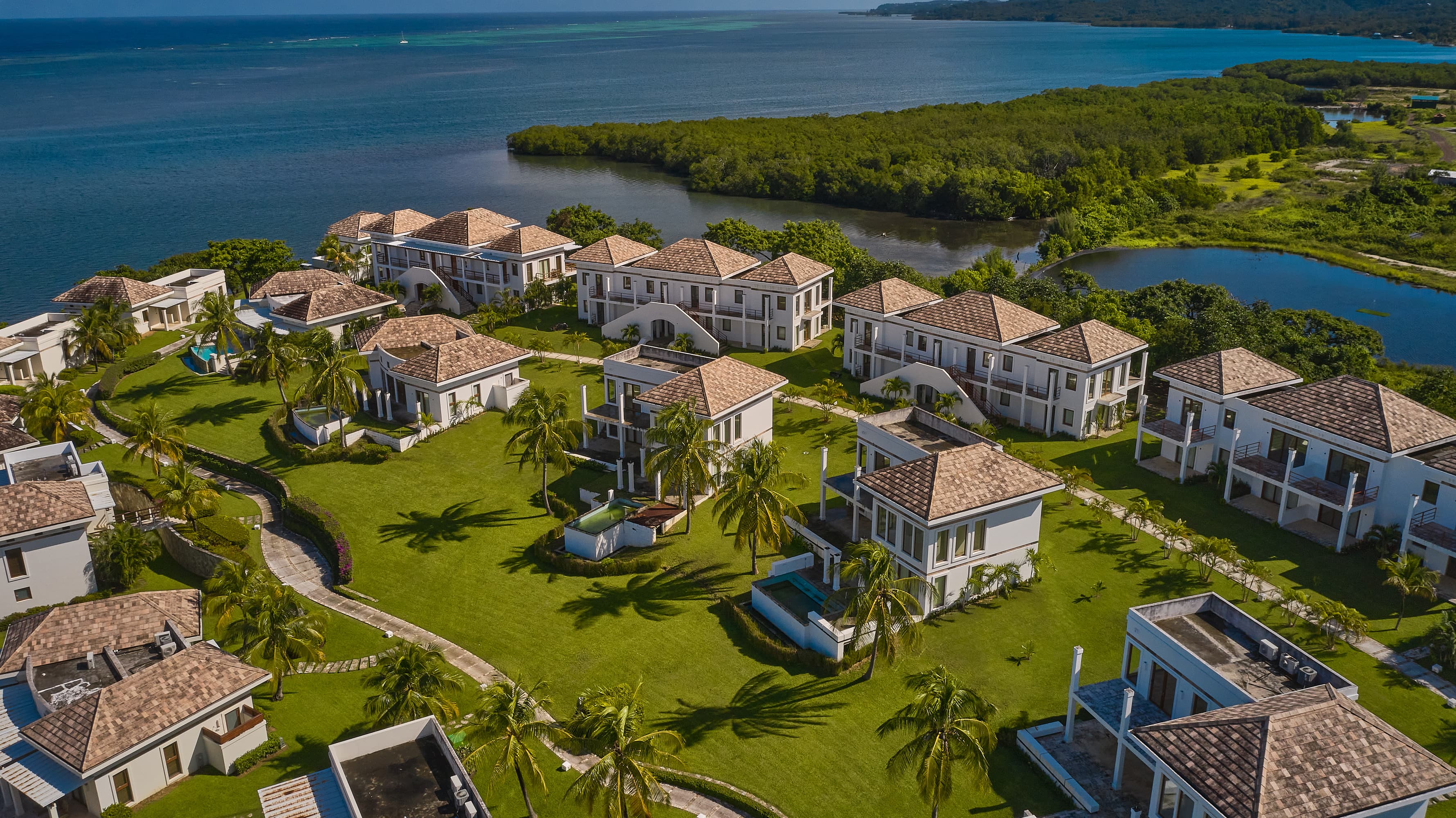 Aerial view of a waterfront resort featuring multiple two-story villas with tiled roofs, each surrounded by well-maintained lawns and palm trees. The villas are situated along a scenic coastline with calm waters and lush greenery, creating a tranquil and luxurious environment.