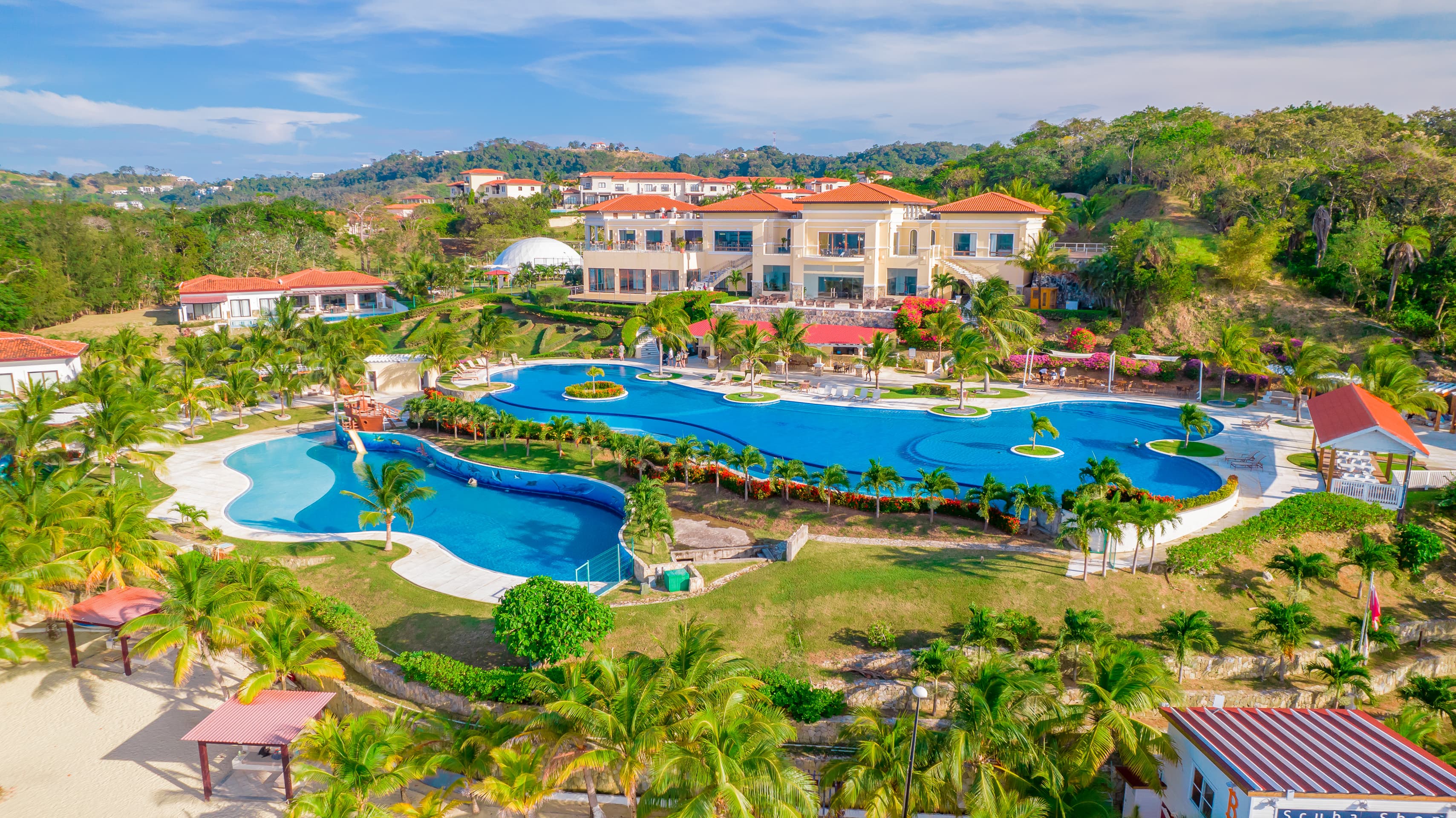 Aerial view of a resort featuring a large, winding swimming pool surrounded by lush greenery and palm trees. The resort buildings have terracotta roofs and overlook the pool area, with hills and additional villas visible in the background.