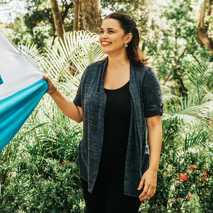 A woman smiling and holding a blue and white flag outdoors, surrounded by lush greenery and trees. She is dressed in a navy cardigan over a black top, creating a cheerful and natural scene.