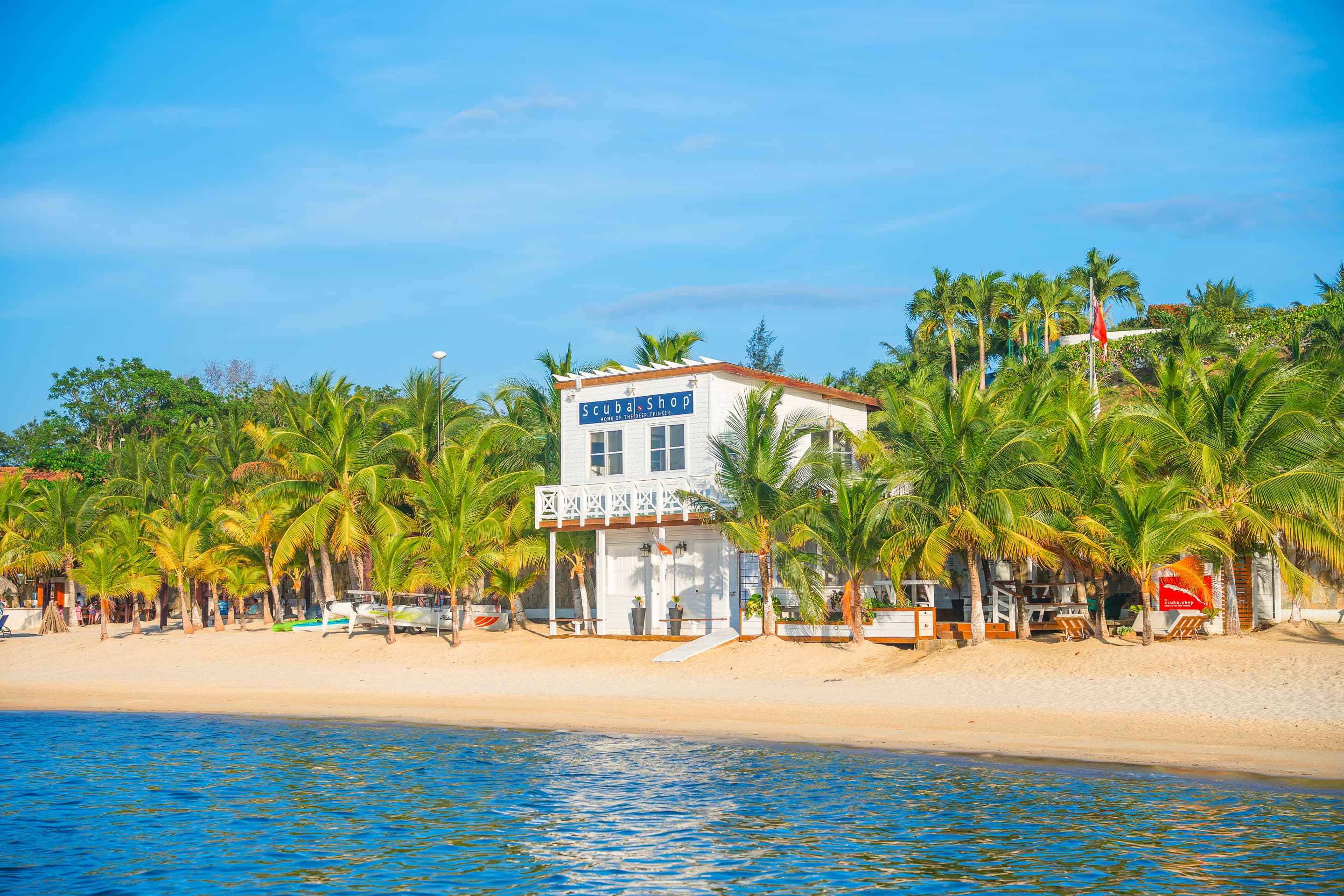 A white two-story building labeled Scuba Shop situated on a sandy beach, surrounded by palm trees. The building faces a calm, blue ocean, creating a picturesque and inviting scene perfect for beachgoers and scuba enthusiasts.