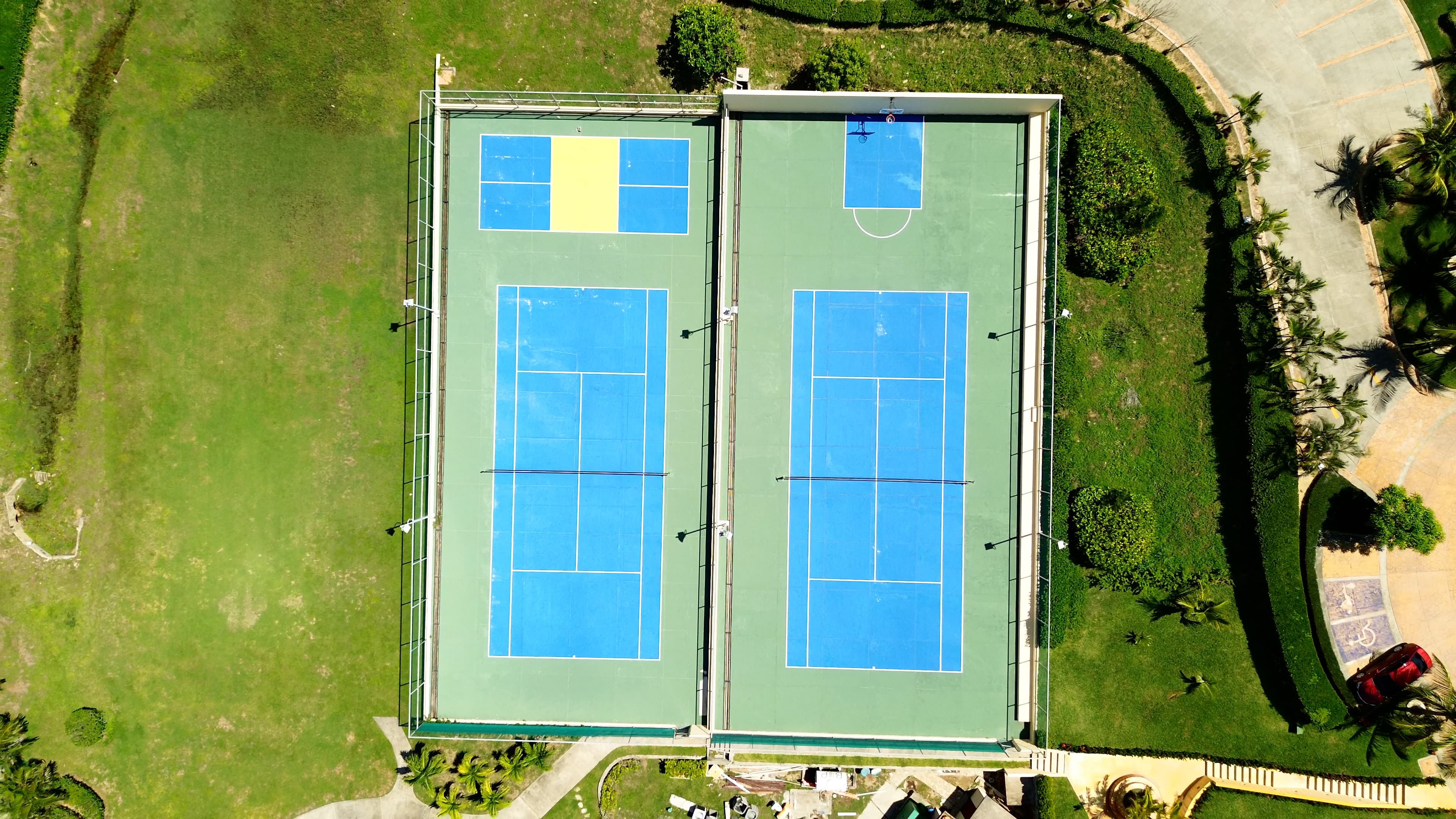 Aerial view of two adjacent tennis courts with blue surfaces, surrounded by green grass and landscaping. One court also features a smaller, yellow-colored area likely used for a different sport or activity. The courts are well-maintained and situated near a parking area and a red car.